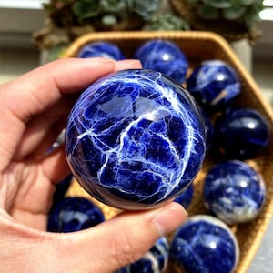 Puede incluir: Un primer plano de una esfera de sodalita azul y blanca. La esfera es lisa y pulida, con un patrón de remolino de azul y blanco.