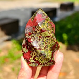 May include: A polished, freeform stone sculpture with a vibrant mix of colors. The stone features a blend of deep red and olive green hues, creating a unique and eye-catching pattern. The sculpture's shape is organic and smooth, with a glossy finish.