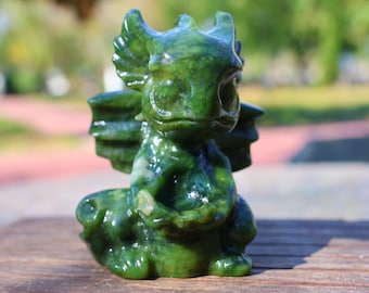 Op de afbeelding: Een gedetailleerd groen jade drakenbeeldje met vleugels en een zittende houding. De draak heeft een gedetailleerd gezicht, hoorns en een getextureerd lichaam. Het beeldje wordt getoond op een houten oppervlak, met een vage buitenachtergrond.