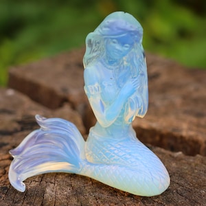 Op de afbeelding: Een doorschijnend, opalescent zeemeerminbeeldje. De zeemeermin heeft lang, vloeiend haar, een gedetailleerde staart en poseert met gevouwen handen. Het beeldje is lichtblauw van kleur en staat op een verweerd houten oppervlak.