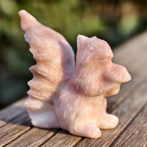 Puede incluir: Una figura de ardilla rosa tallada con una cola tupida y una textura de pelaje detallada. La ardilla está de pie, con las patas delanteras cerca del cuerpo. La figura está hecha de piedra rosa claro.