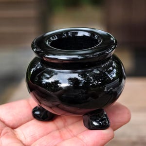 Puede incluir: Un pequeño objeto negro brillante en forma de caldero, con tres patas redondeadas. El caldero tiene una amplia abertura circular y un cuerpo redondeado. La superficie refleja el entorno. El objeto se sostiene en una mano.