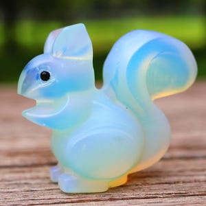 Pode incluir: Uma estatueta de esquilo translúcida e opalescente. A escultura apresenta um corpo azul claro com uma cauda curva e uma base amarelo-alaranjada. O esquilo tem olhos pretos e a boca aberta, dando-lhe uma aparência divertida. A estatueta está sobre uma superfície de madeira.