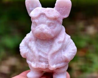 Può includere: Statuina in pietra rosa chiaro di un cane con giacca e cappello. Il cane ha grandi orecchie ed è in piedi. La statuina è scolpita con dettagli ed è tenuta in una mano.