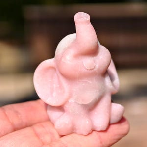 May include: A small, pink elephant figurine carved from stone. The elephant is sitting with its trunk raised and ears spread. The figurine is a light pink color and has a smooth, polished surface.