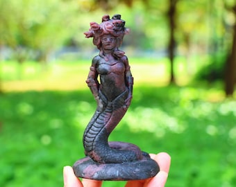 May include: A statue of Medusa, a mythological figure, is depicted. The sculpture is primarily dark with reddish-purple accents. The figure has a woman's torso and a serpent's tail. Her hair is a mass of snakes. The statue is held in a hand.