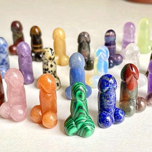 Puede incluir: Una colección de 20 pequeñas esculturas de piedra talladas y pulidas en varios colores y formas. Las esculturas están dispuestas en semicírculo sobre una superficie blanca.