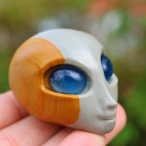 May include: A small, hand-painted ceramic sculpture of an alien head with blue eyes and a brown and white painted design.