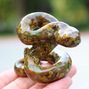 Puede incluir: Una figura decorativa de serpiente hecha de un material brillante y moteado. La escultura presenta un diseño en espiral en tonos marrones, amarillos y verdes. La cabeza de la serpiente está levantada. El fondo está borroso.