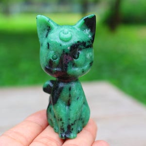 May include: A green and black carved stone cat figurine. The cat has pointed ears, a small nose, and round eyes. The figurine is held in a hand, with a blurred green background.
