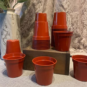 May include: A collection of terracotta-colored plastic plant pots. Some pots are stacked, while others are arranged individually. The pots are cylindrical with a slightly flared rim. A wooden box and a decorative vase are in the background.