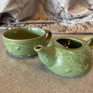 May include: A green ceramic tea set featuring a teapot and a teacup. Both pieces have a textured surface with embossed leaf and vine designs. The set is placed on a neutral-colored surface.