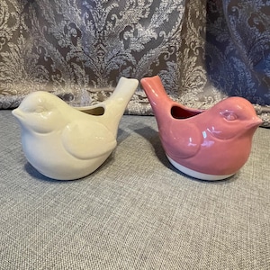 May include: Two ceramic bird-shaped planters. One is cream-colored, the other is pink. Both have a small opening at the top and a white base. The planters are designed to resemble birds.