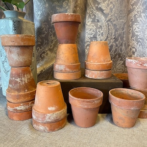 May include: A collection of various terracotta flower pots in different sizes and shapes. Some pots are stacked, showcasing their weathered, aged appearance. The pots are a warm, earthy orange-brown color, with visible signs of use.