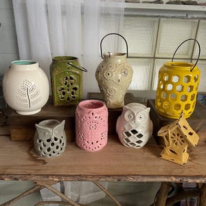 May include: Assortment of ceramic candle holders in various shapes and colors, including owls, houses, and geometric designs. The holders are arranged on a wooden table with a rustic branch base.