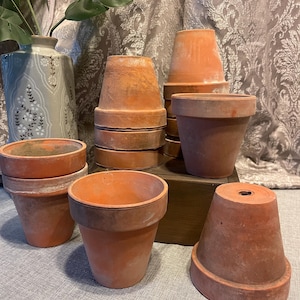May include: A collection of terracotta flower pots in various sizes. The pots are a warm orange-brown color, with a slightly weathered appearance. Some pots are stacked, while others stand alone. The scene includes a decorative vase and greenery in the background.
