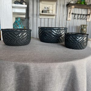 May include: Three dark gray ceramic planters with a geometric diamond pattern. The planters are arranged on a gray tablecloth. The planters are different sizes, with the largest on the left and the smallest on the right. The planters have a glossy finish.