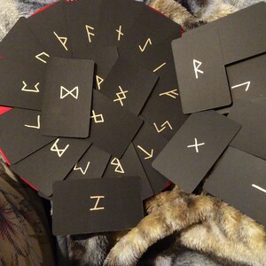 May include: A collection of black rectangular cards with gold-colored runic symbols. The cards are arranged on a red surface, with some cards overlapping. The background is a soft, textured fabric.