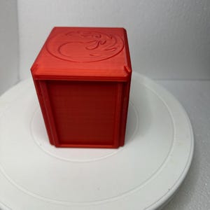 Könnte beinhalten: Eine rote würfelförmige Box mit einem erhabenen, stilisierten Flammenmuster auf der Oberseite.