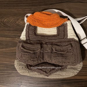 May include: A crocheted brown, white, and orange bag with a white strap. The bag has a pocket on the front and the text "etsy@eleutheromamiayarn" is visible on the bottom.