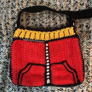 May include: A handmade crochet bag in a vibrant red, yellow, and black color scheme. The bag features a design reminiscent of a jacket, with a pocket detail and a zipper-like pattern down the center. The strap is black, and the bag has a rectangular shape.