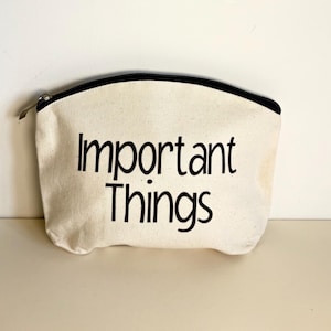 May include: An off-white canvas pouch with a black zip and the words "Important Things" printed in black. The pouch has a black trim along the zip.
