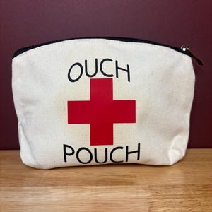May include: Cream canvas pouch with a black zip and the words "OUCH POUCH" printed in black. A large red cross is centered between the words. The pouch is on a wooden surface.