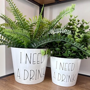 May include: Two white ceramic plant pots with the text "I NEED A DRINK" in black lettering. The pots are filled with green artificial plants. The pots are on a wooden shelf.