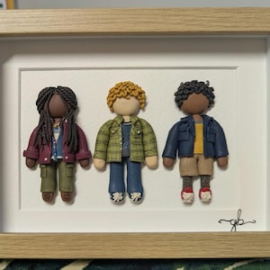 May include: A framed wall art piece featuring three clay figures. The left figure has dark braided hair, a maroon jacket, and olive trousers. The centre figure has curly blonde hair, a green plaid shirt, and blue jeans. The right figure has dark curly hair, a navy jacket, and tan shorts.