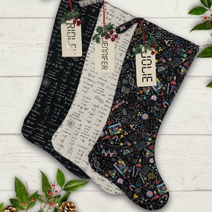 Puede incluir: Tres calcetines navideños con etiquetas con nombres. Los calcetines presentan patrones de temática científica en negro, blanco roto y negro con gráficos coloridos. Cada calcetín está decorado con acebo y bayas.