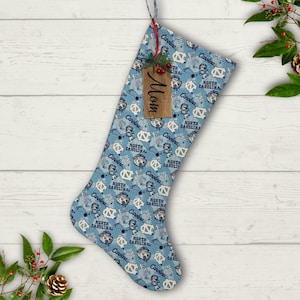 May include: A blue and white Christmas stocking with a repeated pattern of a mascot wearing a hat and the words "North Carolina". The stocking has a wooden tag that says "Mom".