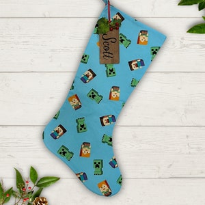 Puede incluir: Un calcetín navideño azul claro con un patrón de personajes pixelados. Una etiqueta de madera con el nombre "Scott" cuelga de la parte superior. El calcetín está decorado con una ramita de bayas y vegetación.