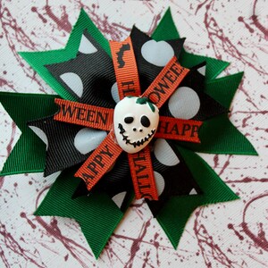 May include: A green, black, and orange Halloween hair bow with white polka dots and a white skull with a black face. The bow has the words "Happy Halloween" and "Ween" on it.
