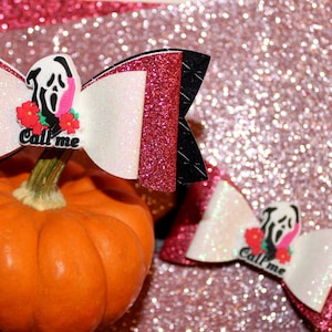 May include: Two Halloween-themed hair bows with a glittery red and black design, featuring a white center with a Scream mask and the text "Call me". One bow is placed on top of an orange pumpkin.