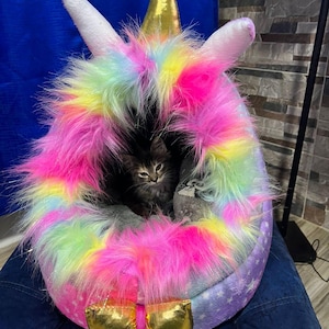 May include: A whimsical cat bed shaped like a unicorn. The bed features a fluffy, rainbow-colored mane and a gold horn and hooves. A small, gray tabby cat is nestled inside the cozy, plush interior.