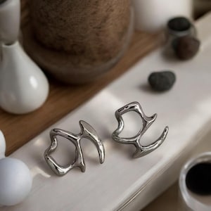 Puede incluir: Dos anillos abstractos de plata con un diseño orgánico en forma de rama. Los anillos están sobre una superficie blanca.