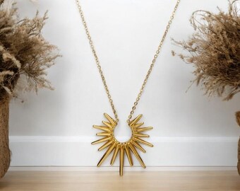 May include: A gold necklace with a large, sun-shaped pendant. The pendant is made of many thin, pointed pieces of metal that radiate outwards from a central circle.