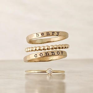 Puede incluir: Cuatro anillos dorados apilados. El anillo superior tiene el nombre "henry" estampado. El segundo anillo tiene un diseño de cuentas. El tercer anillo tiene el nombre "connor" estampado. El anillo inferior tiene un pequeño diamante.