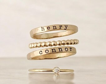 Puede incluir: Cuatro anillos dorados apilados. El anillo superior tiene el nombre "henry" estampado. El segundo anillo tiene un diseño de cuentas. El tercer anillo tiene el nombre "connor" estampado. El anillo inferior tiene un pequeño diamante.