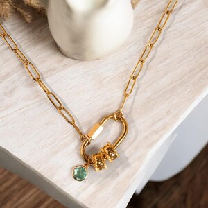 Può includere: Una collana a catena dorata con una chiusura a moschettone dorata e un ciondolo di pietra preziosa verde. La collana ha una serie di piccoli anelli dorati attaccati al moschettone.