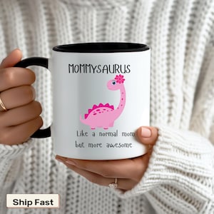 May include: White ceramic coffee mug with a black rim and handle. The mug features a pink dinosaur with a flower on its head and the text "Mommyaurus Like a normal mom but more awesome".