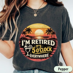 May include: Dark gray t-shirt with a sunset graphic. The graphic includes palm trees, a cocktail, and the text "I'M RETIRED - IT'S 5 O'CLOCK EVERYWHERE" in white and yellow. The shirt is a relaxed fit.