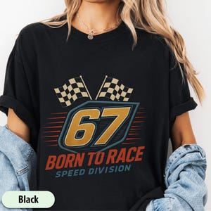 May include: Black t-shirt with a racing-themed design. The shirt features the number 67 in gold, the text "BORN TO RACE SPEED DIVISION", and two checkered flags. The shirt is made of a soft material.