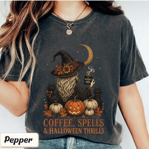 May include: A charcoal gray t-shirt featuring a graphic of a witch wearing a floral hat, holding a mug labeled "WITCH'S BREW" under a crescent moon. The design includes two black cats, pumpkins, and the text "COFFEE, SPELLS & HALLOWEEN THRILLS."