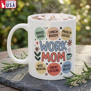 May include: White ceramic mug with the text "WORK MOM" in colorful letters, surrounded by phrases like "Advice Giver" and "Office Bestie." The mug is filled with hot chocolate and marshmallows. "MADE IN THE USA" is printed on the top left.