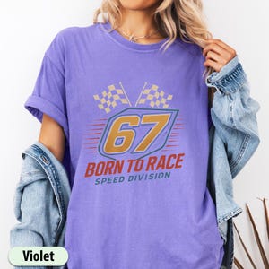 May include: Violet-colored t-shirt with a racing-themed graphic. The design features the number 67 in gold, the words "BORN TO RACE" in red, and two checkered flags. The shirt also includes the text "SPEED DIVISION".