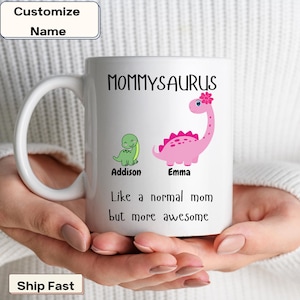 May include: White ceramic coffee mug with a pink and green dinosaur design. The text "Mommyaurus" is printed on the mug, along with the text "Like a normal mom but more awesome".