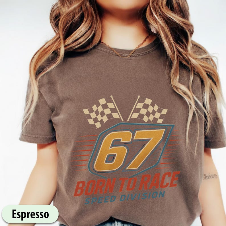 May include: Espresso-colored t-shirt with a racing-themed graphic. The design features the number "67" in gold, two checkered flags, and the text "BORN TO RACE SPEED DIVISION" in red and blue.