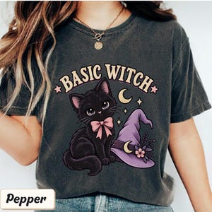 May include: A charcoal gray t-shirt featuring a graphic of a black kitten with a pink bow, a purple witch hat with a crescent moon and flower, and the text BASIC WITCH in a curved font. The design includes small stars and a crescent moon.