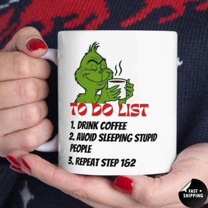 May include: White ceramic mug featuring a green cartoon character holding a coffee cup. The mug has the text "TO DO LIST" followed by three steps: drink coffee, avoid sleeping stupid people, and repeat steps 1 & 2.